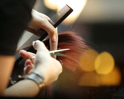 hands holding scissors and comb cutting long red hair in salon setting with blurred background showing beauty and hair styling techniques